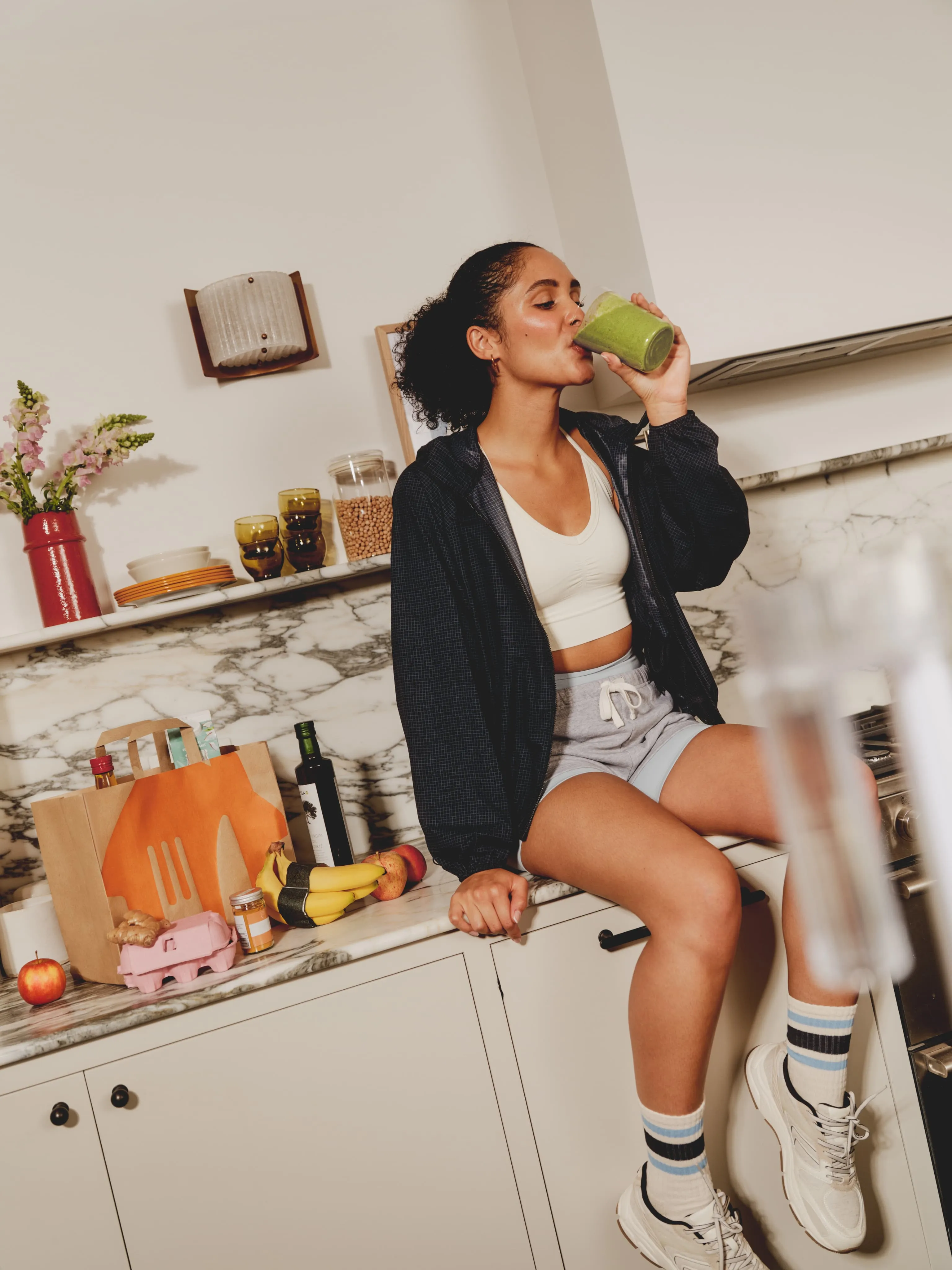 kobieta w sportowym ubraniu pije zielone smoothie siedząc na kuchennej szafce, obok niej widoczna jest siatka z dostawy Pyszne.pl