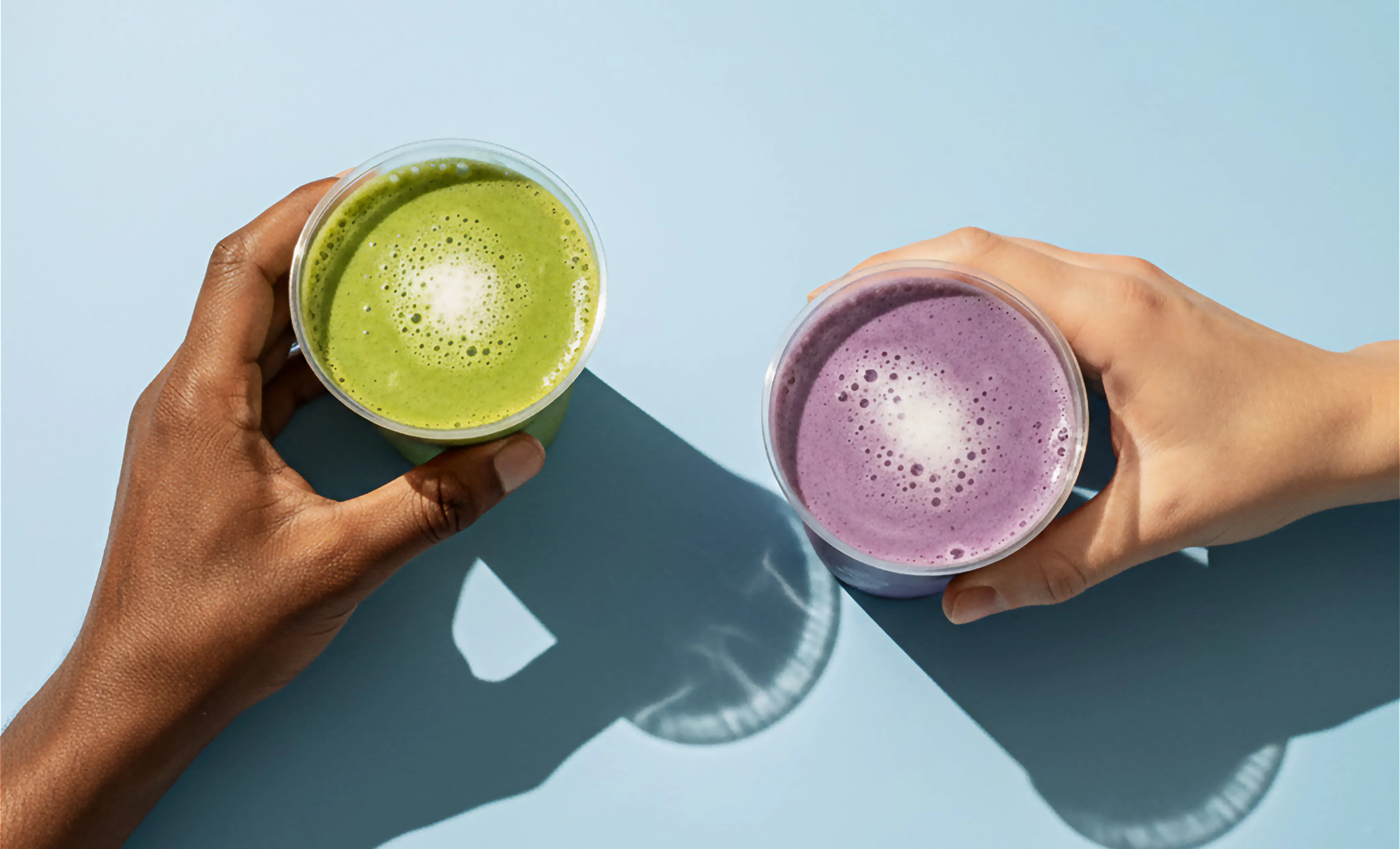  dwie dłonie, każda trzymająca swój kubek z napojem - w jednym jest zielona, a w drugim fioletowa matcha
