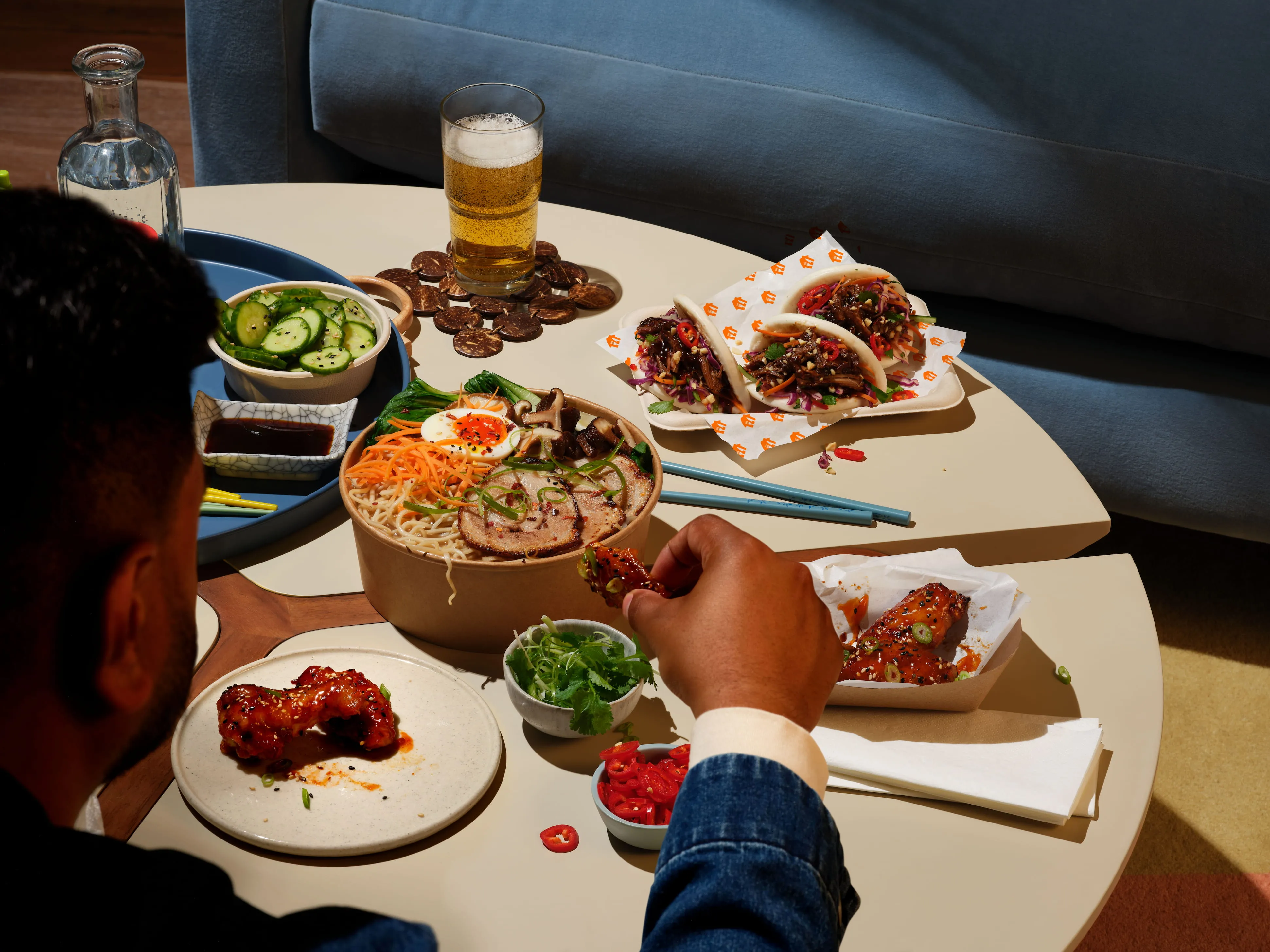 Stół z miską ramen, ogórkami, taco z mięsem, piwem oraz skrzydełkami na niskim stoliku, a osoba sięga po jedno z nich.