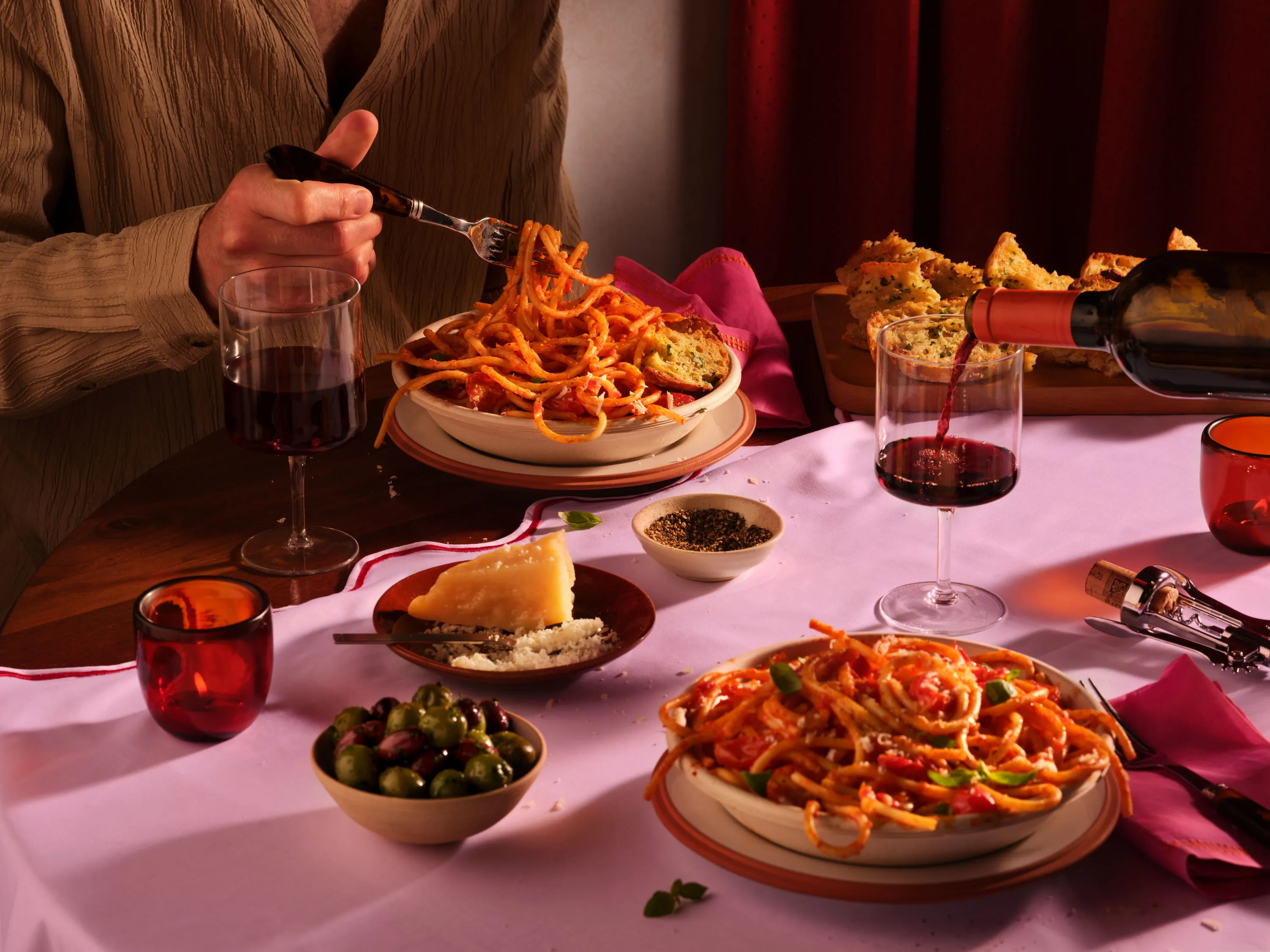 Stół z talerzami spaghetti, kieliszkami czerwonego wina, miseczką oliwek, kawałkiem sera i pieczywem, z ręką sięgającą po makaron.